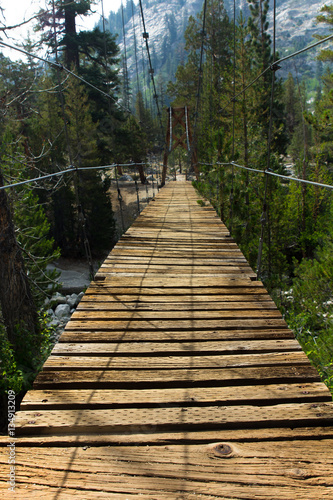 An old wooden suspension bridge with broken slats hangs over a river near the Rae Lakes in California's high sierra
