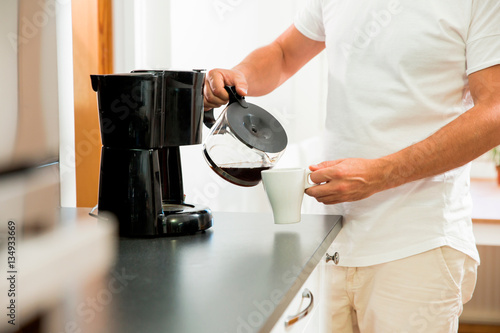 Obraz na plátně Man in the kitchen pouring a mug of hot filtered coffee from a glass pot