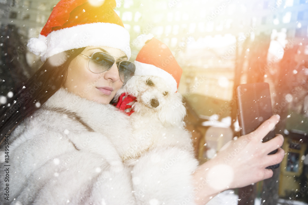 Beautiful young woman with her dog. Wearing Christmas hats. Smiling young woman holding her dog while taking a selfie. Lens flare in a background. Beautiful young woman with her dog. Wearing Christmas hats. Smiling young woman holding her dog while taking a selfie. Lens flare in a background.