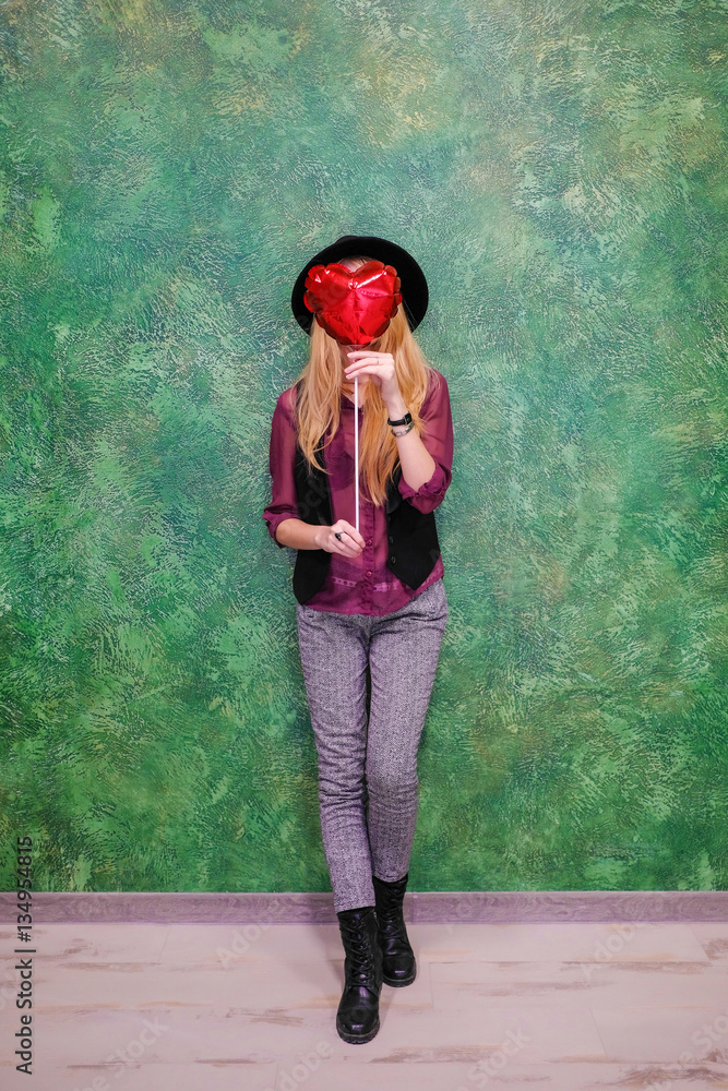 Obraz premium Closeup portrait girl with red balloons with heart shape on a green background. Vertical shot. Girl hipster in a black bowler hat holding a red inflatable heart and celebrate Valentine's day.