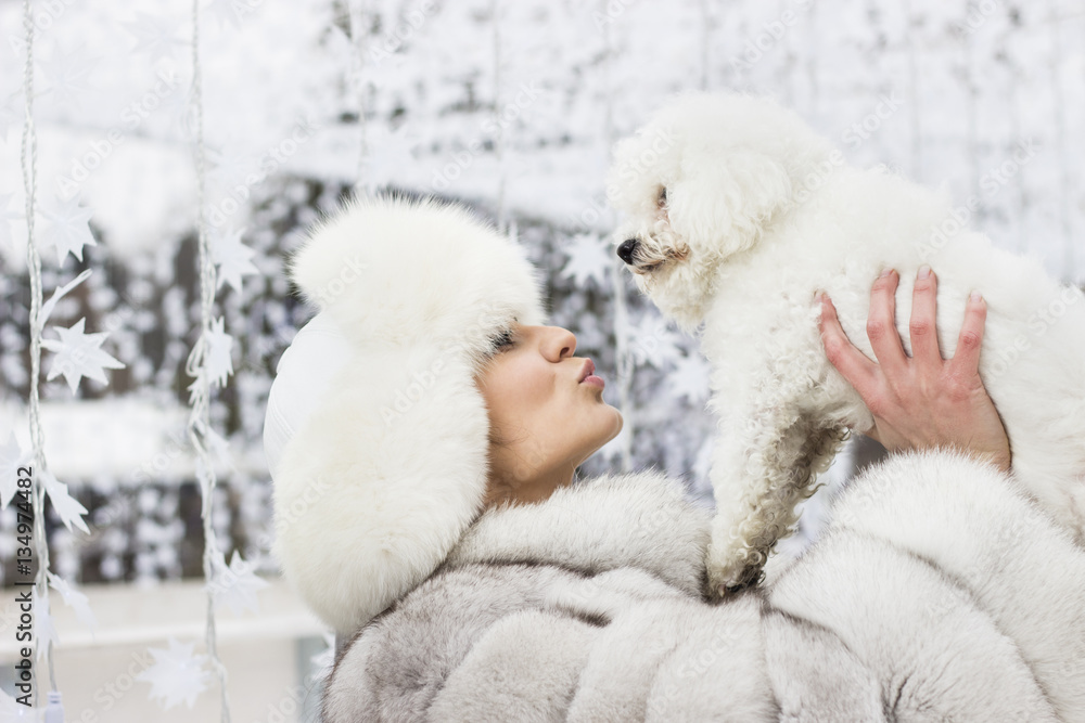 Winter beauty. Close-up of a young woman in the wintertime with her dog. Woman wearing white winter fur coat while holding her dog. Christmas decorations in the background. Winter beauty. Close-up of a young woman in the wintertime with her dog. Woman wearing white winter fur coat while holding her dog. Christmas decorations in the background.