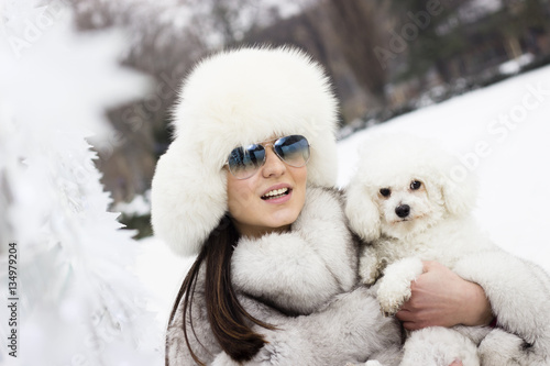 Play time at the park. Woman with winter sunglasses playing with her dog. Young beautiful woman wearing a fur hat and a winter coat.