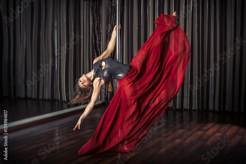 Young pole dancer girl in mask and red skirt sitting indoor near the pole on curtain background