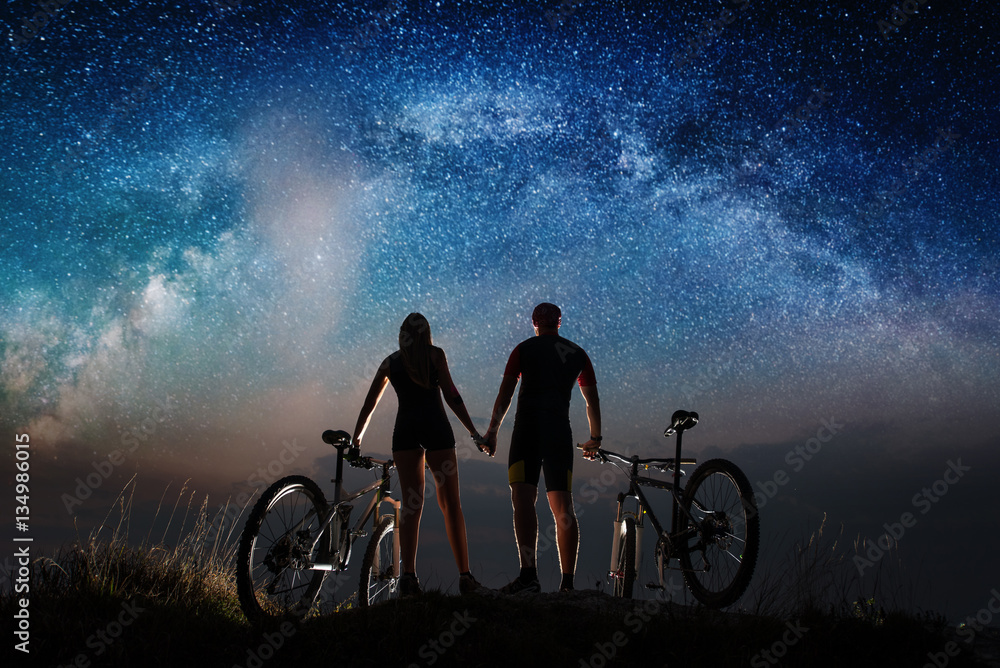 Obraz premium Rear view of silhouette couple cyclists holding hands enjoying night starry sky. Bikers with mountain bicycles on the hill. Night landscape with colorful Milky Way