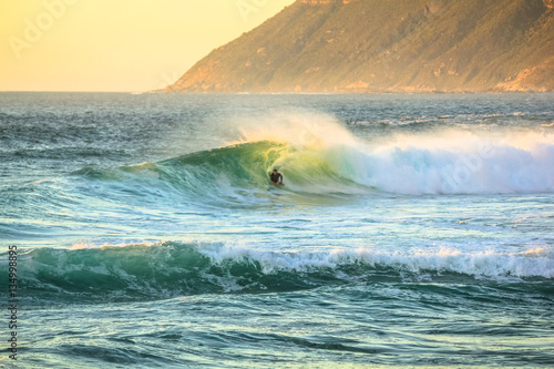 Noordhoek Beach at sunset. Surfing in Cape Town, South Africa. Surfer tries to take a big wave. Atlantic coast in Table Mountain National Park. Extreme sports leisure concept.