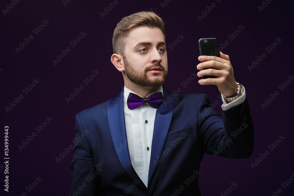 Naklejka premium handsome man in tuxedo and bow tie makes selfie against dark background. Fashionable, festive clothing