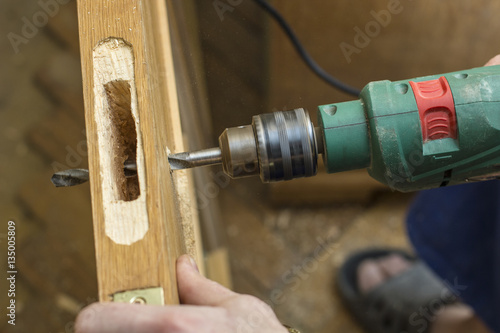 Carpenter makes a hole in old wooden door the mortise lock for by using power drill. Close-up.