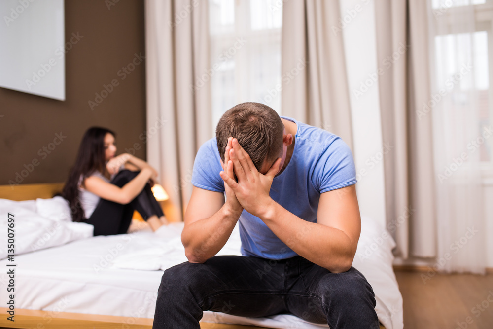 Young couple having a problem. Handsome man is leaning on his hand while sitting sadly on bed, woman is lying in the background Young couple having a problem. Handsome man is leaning on his hand while sitting sadly on bed, woman is lying in the background