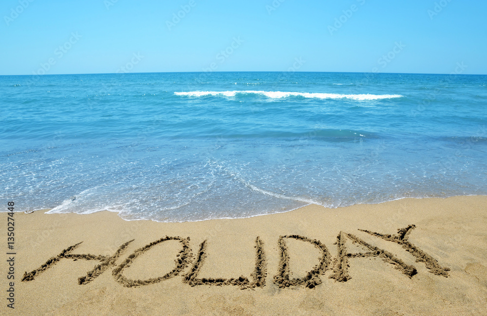Fototapeta premium Holiday written on sandy beach, in the background sea and blue sky.