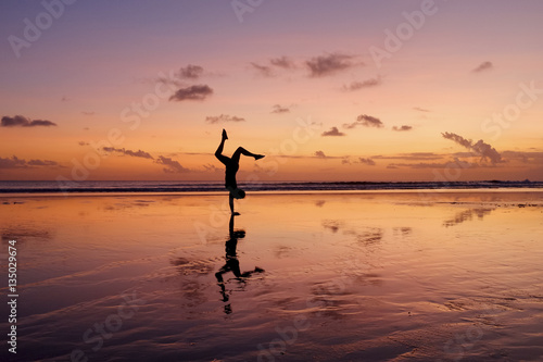A silhouette of a girl and her reflection doing a handstand on a beach during sunset.