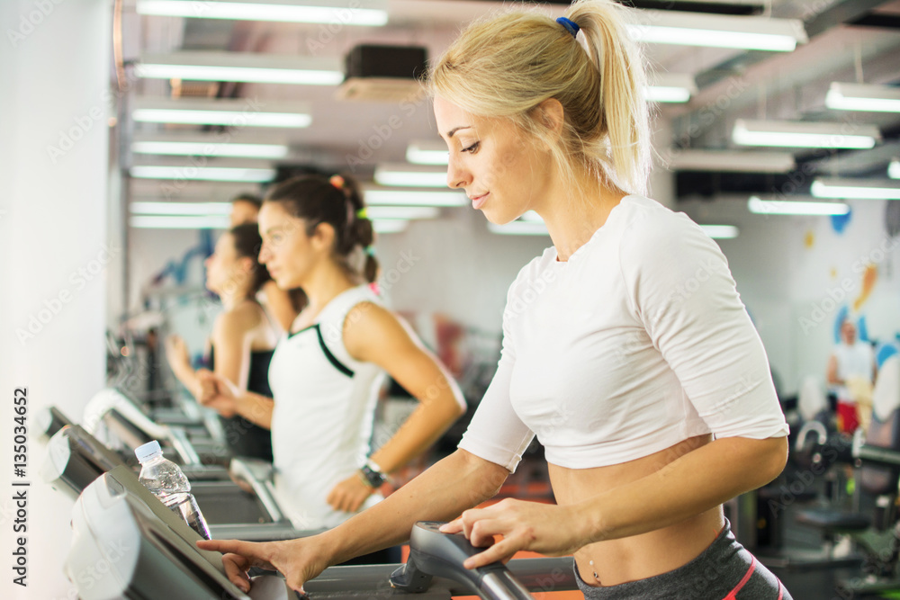 Young female athlete exercising on treadmill in a gym. She is setting up difficulty level on gym machine.