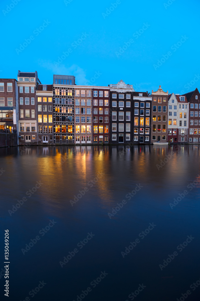 Houses facades over canal with reflections illuminated at blue night, Amsterdam, Netherlands