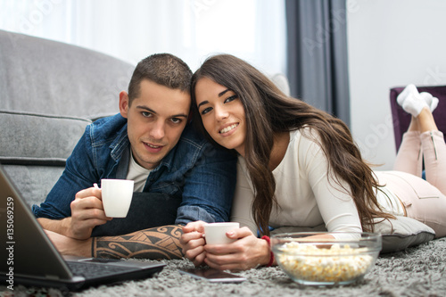 Young couple having coffee cups while lying down on the floor and looking at camera.