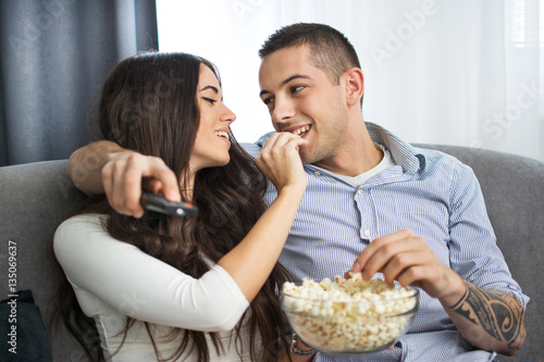 Young woman feeding her boyfriend with popcorn. They are sitting on sofa and watching tv.