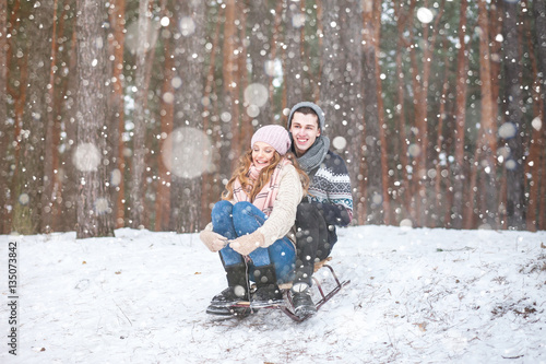 Young couple in winter forest sledding, laughing during snowfall