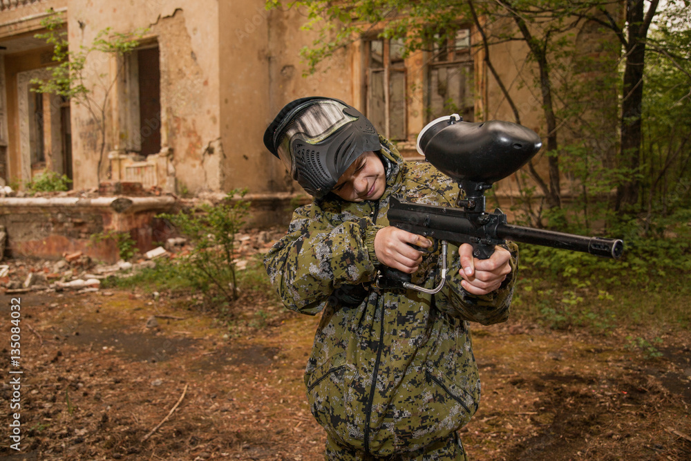 Naklejka premium young boy playing paintball. boy aiming a paintball gun. Boy in camouflage clothing. boy in a protective suit. Boy in a protective mask. game of war. war game. Boy shoots a gun.