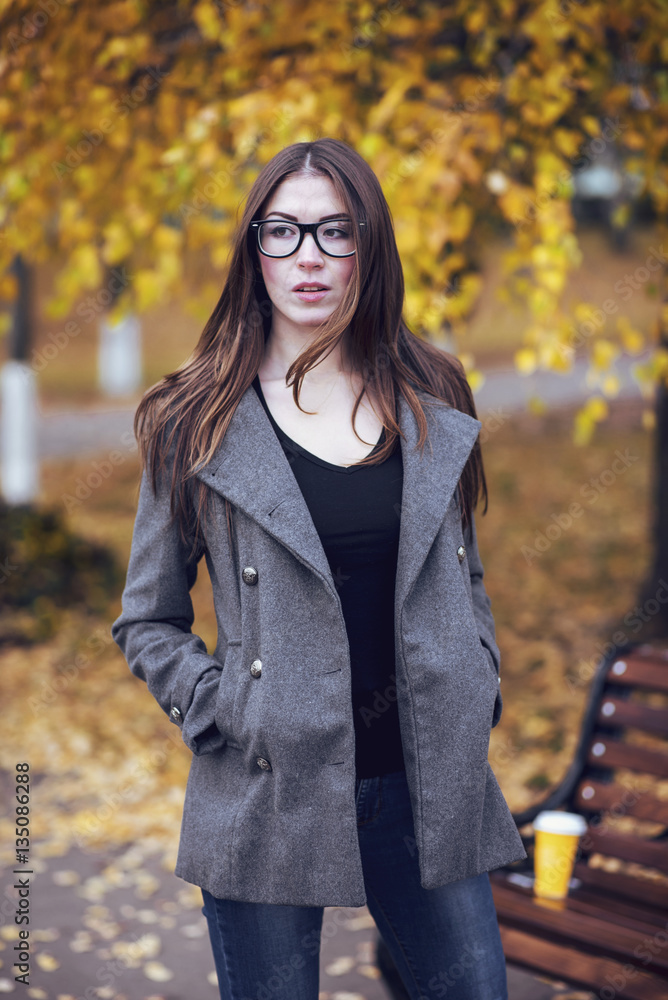 Girl brunette in a park in coat wearing glasses, autumn or spring, walks in the city, waiting for girlfriends. Young long-haired, hipster still life. Girl brunette in a park in coat wearing glasses, autumn or spring, walks in the city, waiting for girlfriends. Young long-haired, hipster still life.