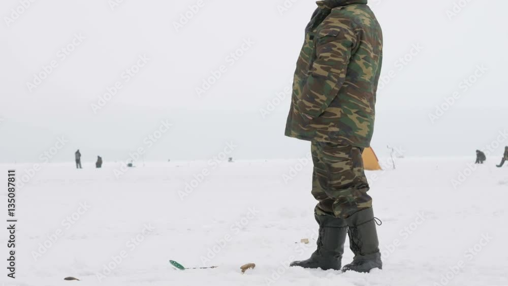 The man is engaged in winter fishing. It costs about fishing rods waiting fish. Warmly dressed in protective clothing The man is engaged in winter fishing. It costs about fishing rods waiting fish. Warmly dressed in protective clothing