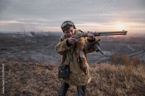 young boy standing on a mountain with a gun. post-apocalyptic world. boy with fox skin on his shoulders. boy aiming a gun. teenager forest hunter. Apocalypse