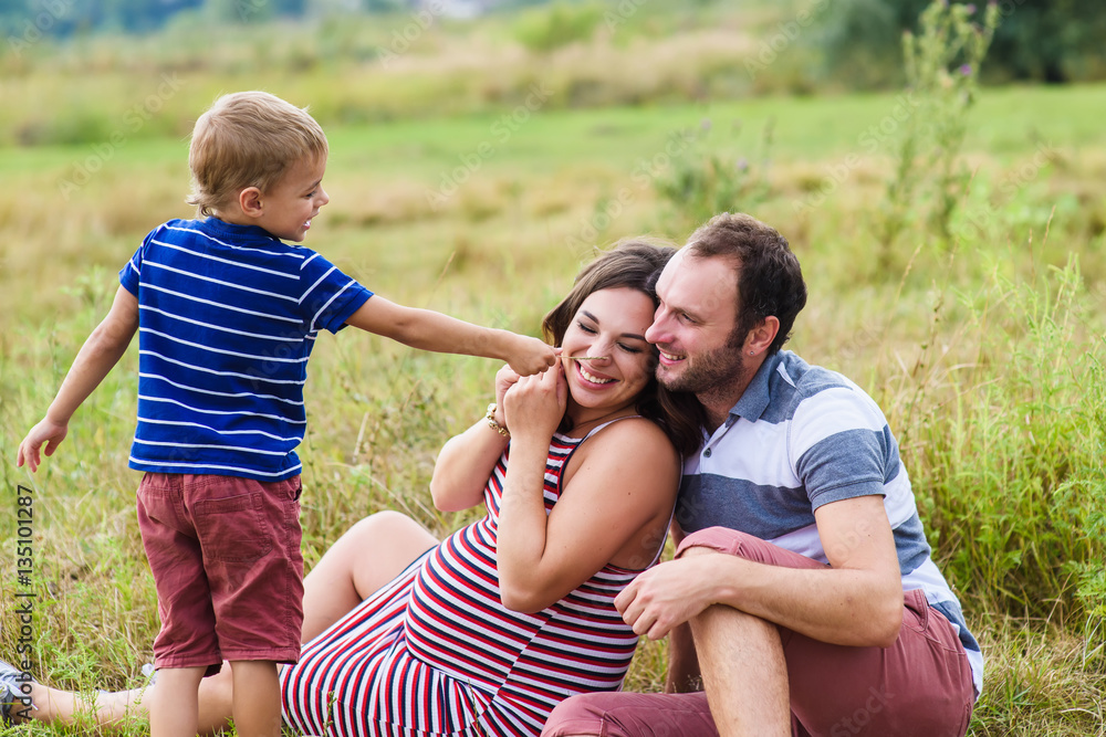 Child tickles his pregnant mother a blade of grass. Mother is expecting a baby. Pregnant woman, husband and their son playing outdoors. Child tickles his pregnant mother a blade of grass. Mother is expecting a baby. Pregnant woman, husband and their son playing outdoors.