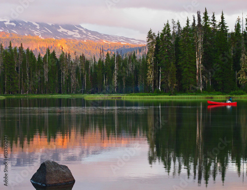 Vászonkép A bright red canoe on a high mountain lake with the forest and mountains reflecting on the calm waters and the orange glow of sunset on a peaceful summer evening