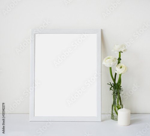Empty white frame with place for text on the white wall and table, bouquet of flowers ranunculus and candle. Scandinavian style room interior. Template mock up for paintings or photographs