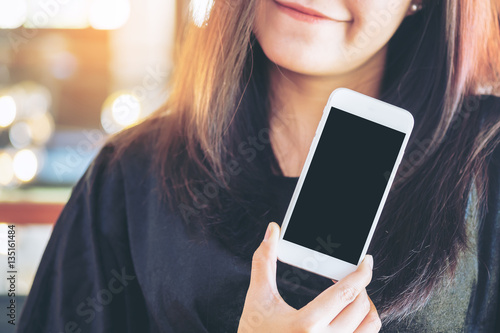 Mockup image of a beautiful woman holding and showing white mobile phone with blank black screen and smiley face in vintage cafe