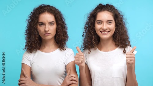 Two beautiful female twins with curly hair standing on blue background. One is grumpy while another showing joy photo