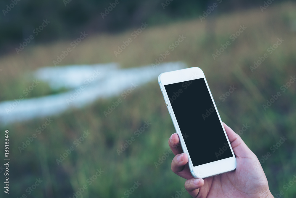 Mockup image of a hand holding and using white mobile phone with blank black screen in green garden nature 