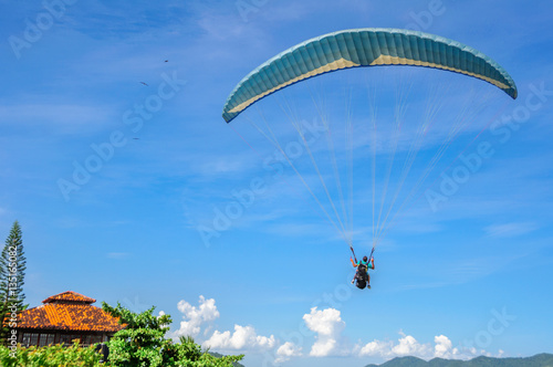 Back of the paragliding man in blue sky with white clouds, green trees and residential house with terracotta tiles and mountains on the background at Camboinhas Beach, Niteroi, Rio de Janeiro, Brazil