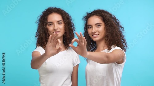 Two beautiful female curly Caucasian twins in white t-shirts synchronously showing ok on blue background in slowmotion photo