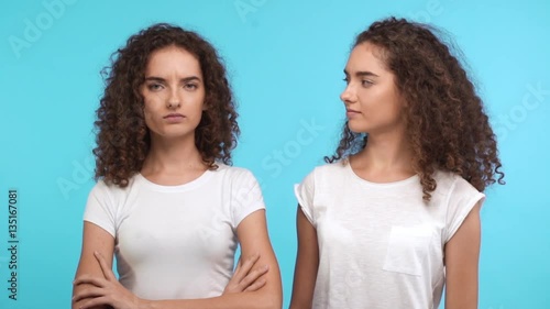 One dissapointed female curly Caucasian twin standing on blue background while another smiling claping hands and kissing her cheek in slowmotion photo