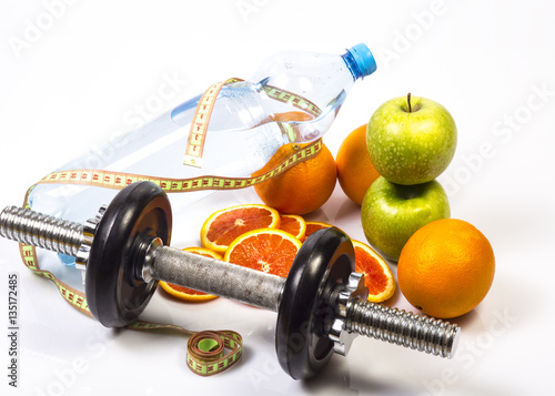 Concept of healthy active lifestyle. Heap of green and red apples and red oranges. Bottle of pure water with flexible ruler. Fitness dumbbell in foreground. Sliced red orange.