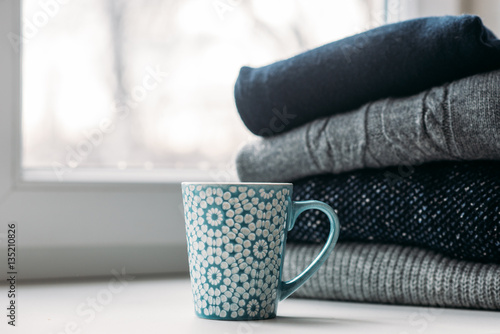 Cozy winter still life scene: a cup, and stack of knitted jumpers and sweaters on window sill