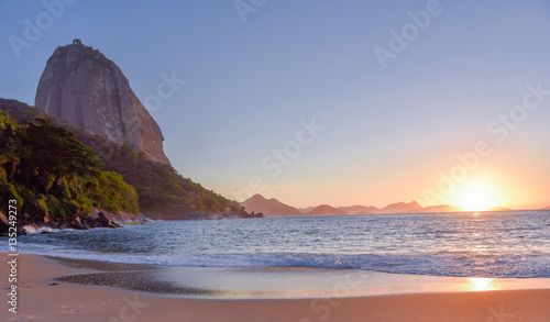 Beautiful sunrise at the deserted Praia Vermelha Beach with the bright sun rising from the Atlantic Ocean, solar path on the water and the Sugarloaf Mountain, Rio de Janeiro, Brazil
