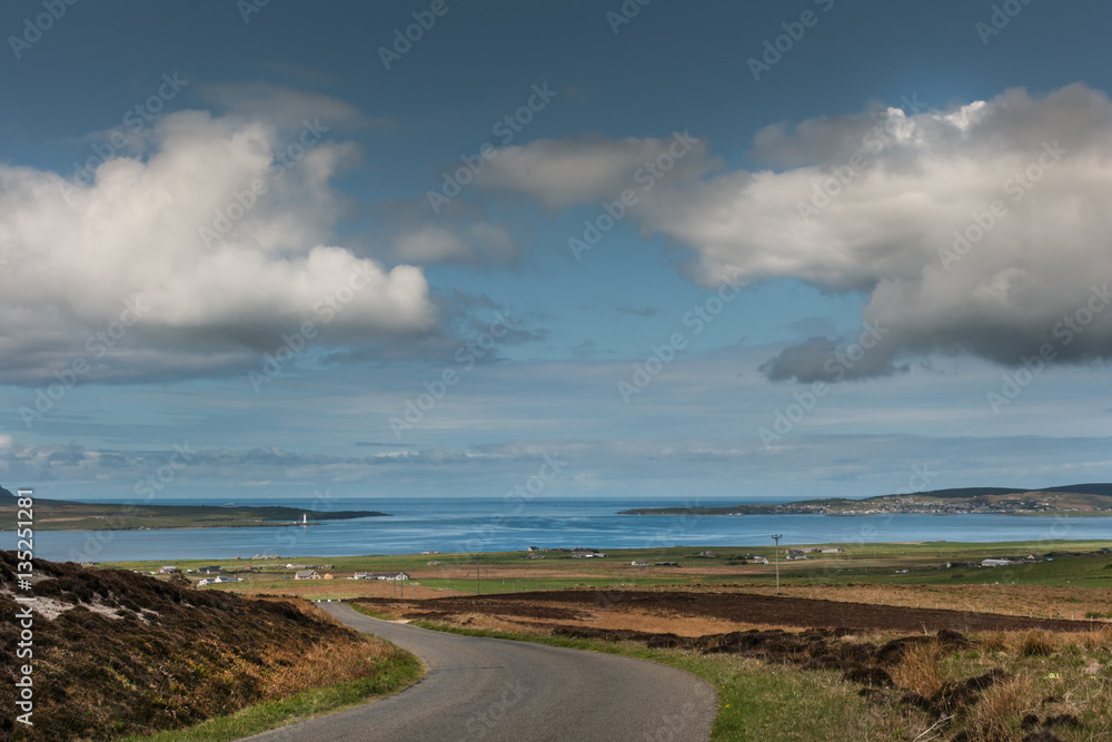Orkneys, Scotland - June 5, 2012: Landscape looking at entrance to blue Atlantic Ocean under blue cloudy sky. Foreground is meandering road between brown fields, green meadows and farms. Main Island. Orkneys, Scotland - June 5, 2012: Landscape looking at entrance to blue Atlantic Ocean under blue cloudy sky. Foreground is meandering road between brown fields, green meadows and farms. Main Island.