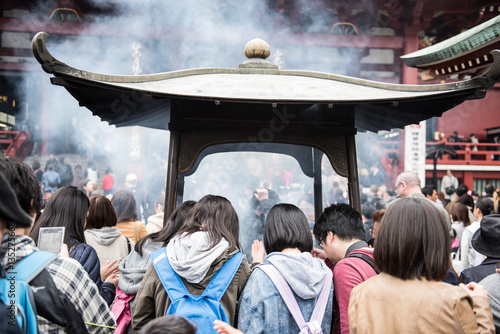 In the picture we can see lots of people praying inside a temple. Smoke is coming out from a chamber where many people are burning incense sticks.