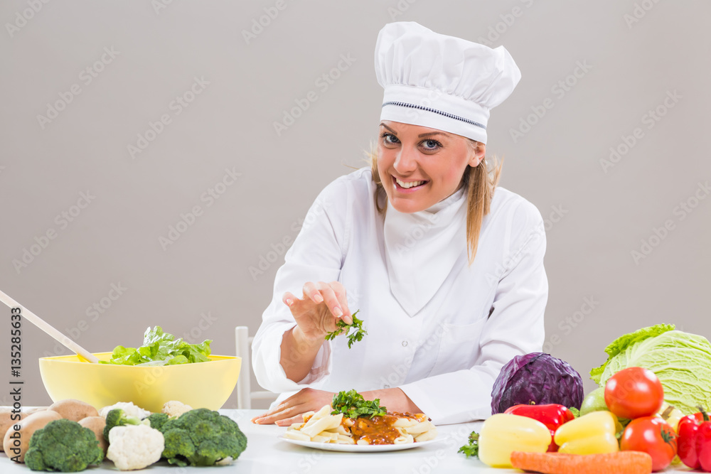 Cheerful female chef is sitting at the table with bunch of vegetable and decorating prepared meal.
Cheerful female chef is sitting at the table with bunch of vegetable and decorating prepared meal.