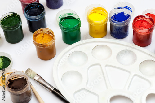Open gouache jars with empty white plastic palette and few paintbrushes on a white background