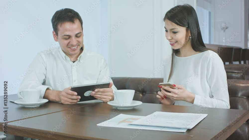 Smiling young man showing something on his tablet to female coworker holding phone during lunch Smiling young man showing something on his tablet to female coworker holding phone during lunch