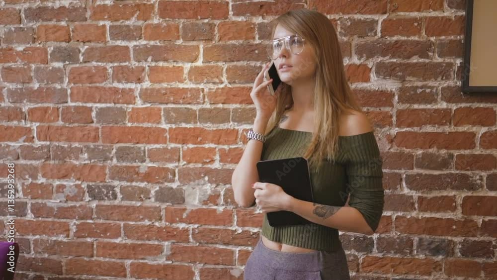 Young cute woman in glasses, standing in room near brick wall and talking on cellphone while holding tablet computer in her hand. Young cute woman in glasses, standing in room near brick wall and talking on cellphone while holding tablet computer in her hand.