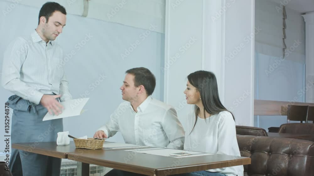 Waiter bringing menu to young couple sitting at the table in a restaurant Waiter bringing menu to young couple sitting at the table in a restaurant