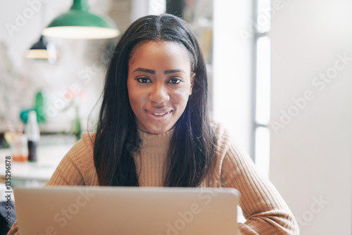 Portrait of attractive african female looking at the camera while sitting with the portable computer in a modern coffee shop. Young entrepreneur is sharing ideas with the partners by the emails.