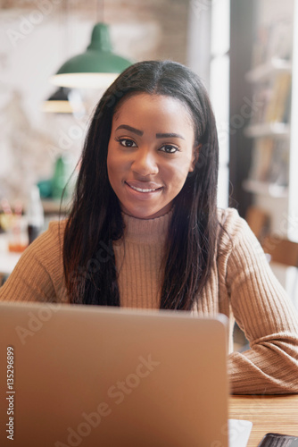 Young beautiful hipster girl is sitting with a latop in a cafe. Attractive african woman with long dark hair is working on a portable computer. A beauty blogger is typing messages on a computer.
