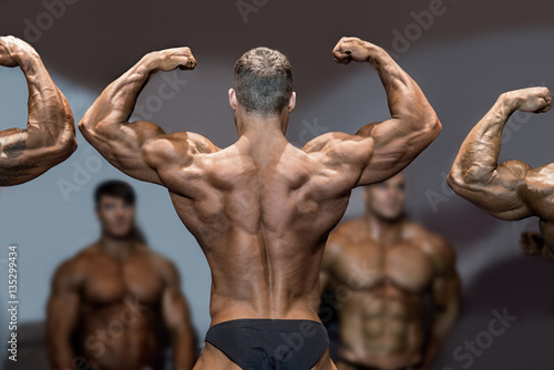 Bodybuilder flexing back and biceps. Muscular athlete posing on stage. Top sportsmen at bodybuilding competition. Opponents are watching.