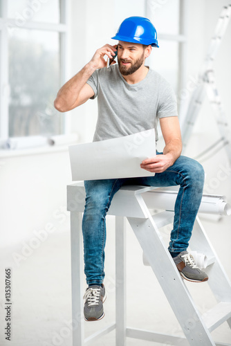 Handsome repairman or builder in helmet working with drawings and phone on the renovation of apartment interior