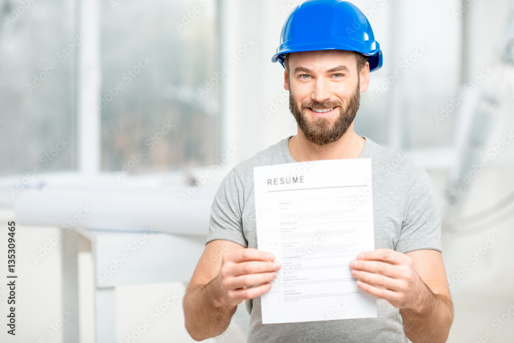 Handsome builder or worker in protective helmet holding his resume standing in the white interior. Handsome builder or worker in protective helmet holding his resume standing in the white interior.