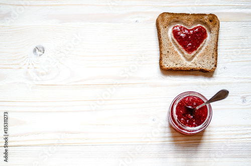 Toasted white bread with a heart inside. At the heart plastered with raspberry jam. Under the croutons is a jar of jam, a jar of sticks dessert spoon.