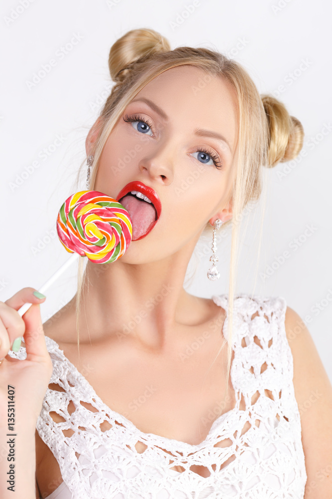 Beauty Model smiling teen girl, licking a lollipop, funny blonde hairstyle - two bumps, professional makeup. Snow-white teeth, blue eyes, long eyelashes. Isolated on white background