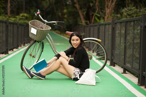 Pretty Thai girl reading book and sitting near by bicycle at the park